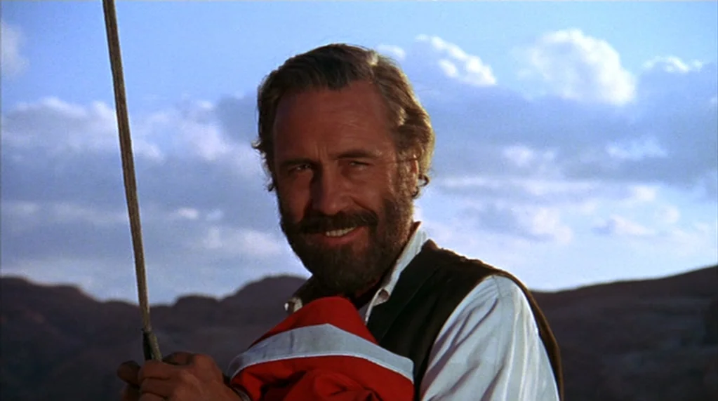 The Ballad of Cable Hogue (1970) movie still: medium — A man stands outdoors under a clear sky with distant arid mountains visible. He holds a red…