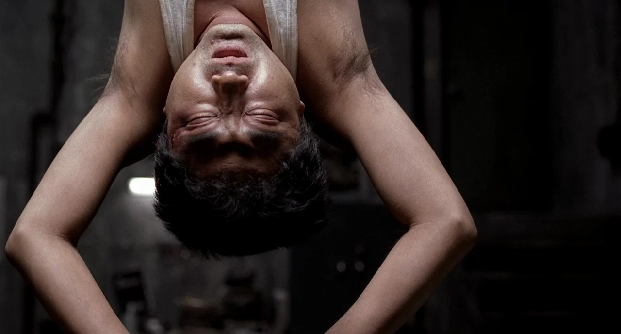 Memories of Murder (2003) movie still: closeup — A dark, industrial-style basement with concrete walls serves as the backdrop. A man hangs upside…