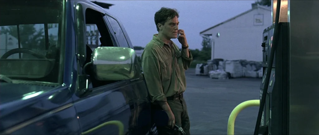 Take Shelter (2011) movie still: medium — An asphalt forecourt during the blue hour at a commercial refueling station. A man stands beside a blue…