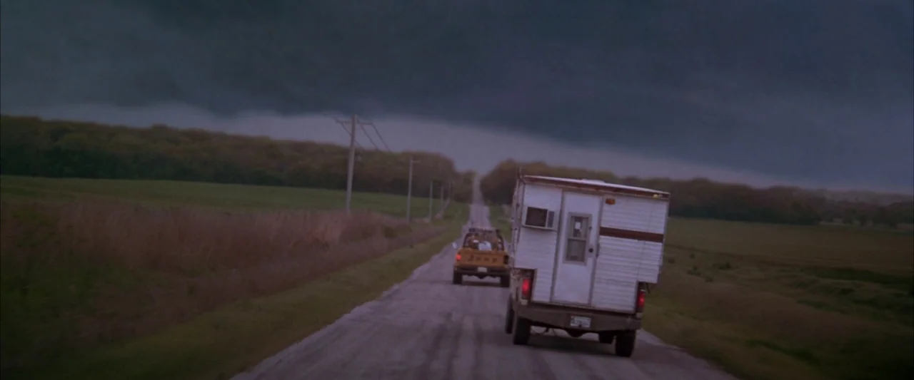 Twister (1996) movie still: wide — A rural asphalt road stretches toward the horizon under a massive, dark, low-hanging storm cloud. A white camper…