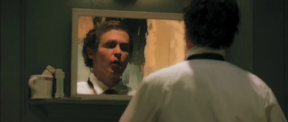 Confessions of a Dangerous Mind (2002) movie still: medium — A dimly lit room with dark green walls features a small rectangular wall mirror. A man…