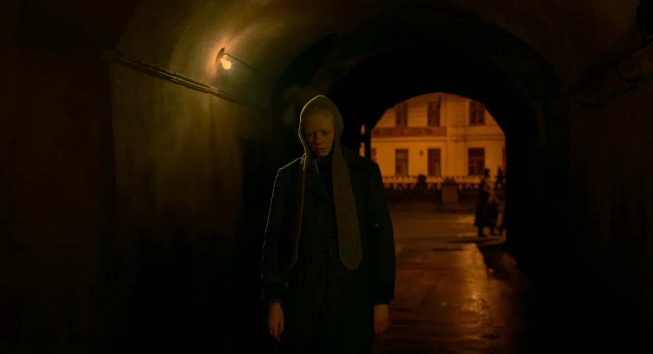 Beanpole (2019) movie still: medium — A person stands in a dimly lit, arched stone tunnel at night. Beyond the tunnel opening, a bright streetscape…