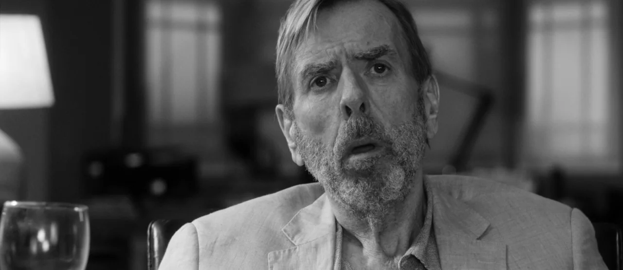 The Party (2017) movie still: closeup — A dimly lit room with an office desk and a glass in the foreground. An elderly man with a beard sits at the…