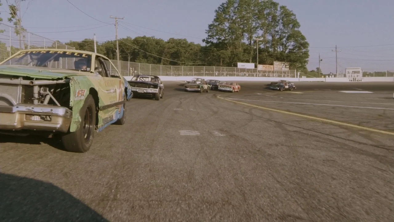 The Last Race (2022) movie still: wide — An asphalt racetrack features a series of stock cars navigating a sharp curve. The surrounding area…