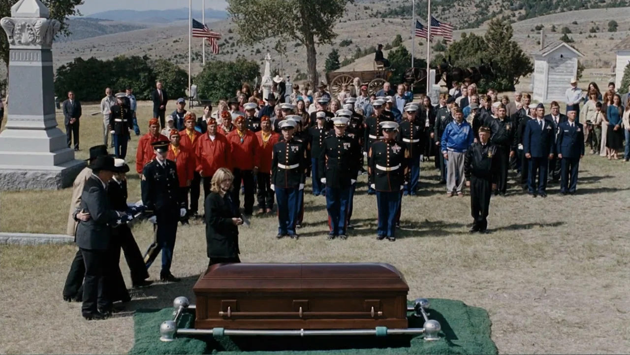 Taking Chances (2011) movie still: wide — An outdoor cemetery setting features a wooden casket resting on a green platform. A large group of…