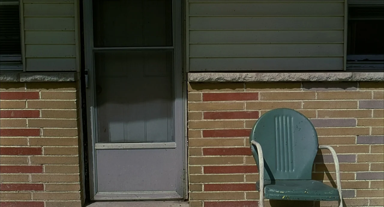 A History of Violence (2005) movie still: wide — An exterior wall of a brick house features a grey screen door and beige siding. A single green…