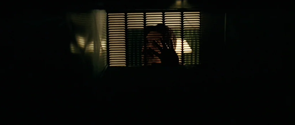 No Country For Old Men movie still: medium — A person looks through horizontal window blinds in a dark room. The subject holds their hand up to the…