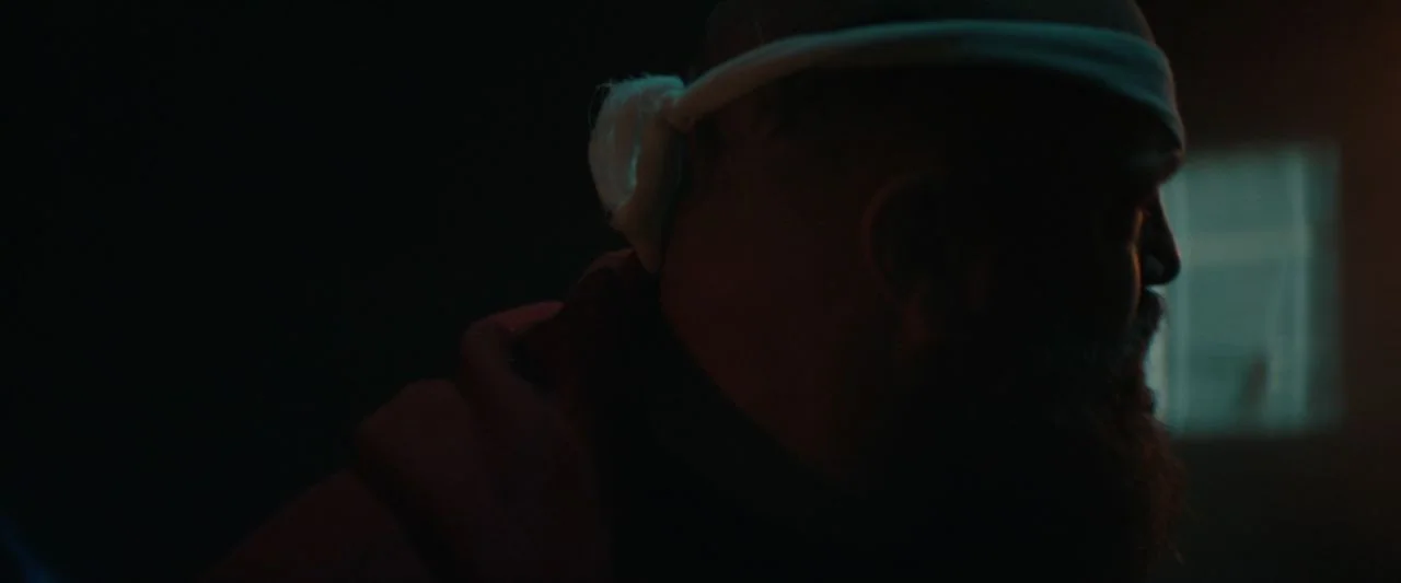 Becky (2020) movie still: closeup — A dark, minimally lit room features a blurred square light source in the background. A man with a dark beard and…