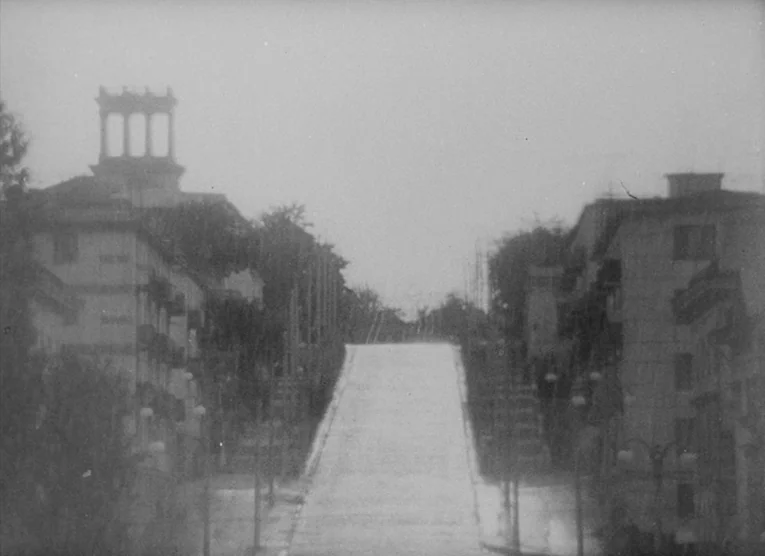 Wings (1966) movie still: establishing — A quiet urban street slopes upward between two rows of classical apartment buildings. Tall streetlights…