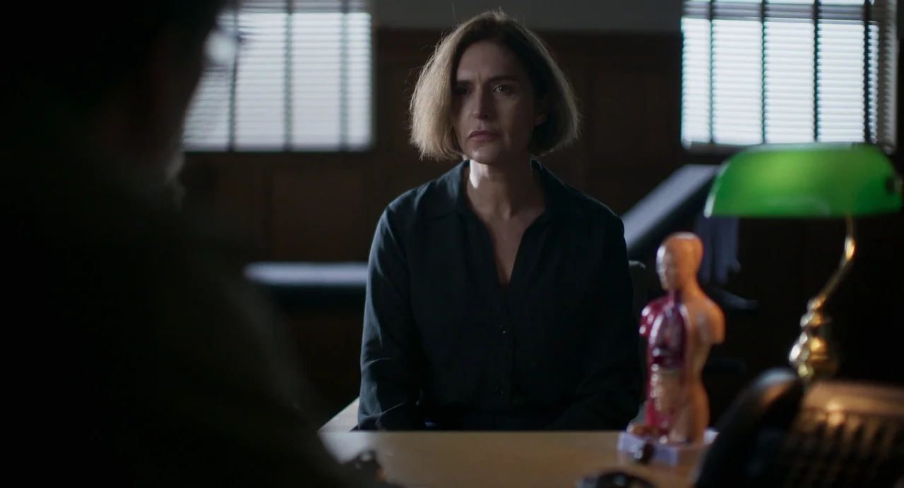 All You Need Is Death (2023) movie still: medium — A quiet office contains a wooden desk with a small anatomical torso model on it. A woman sits…