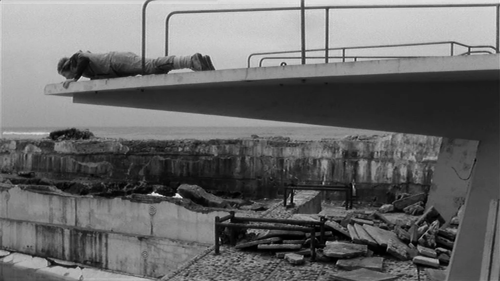 The State of Things (1982) movie still: wide — A desolate, abandoned seaside pool with broken concrete and scattered rubble. A lone individual lies…