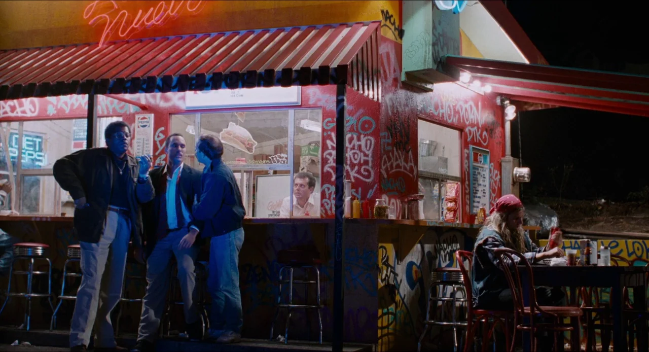 Deep Cover (1992) movie still: wide — A roadside fast food stand covered in heavy graffiti is lit by red neon lights. Several men stand at the order…