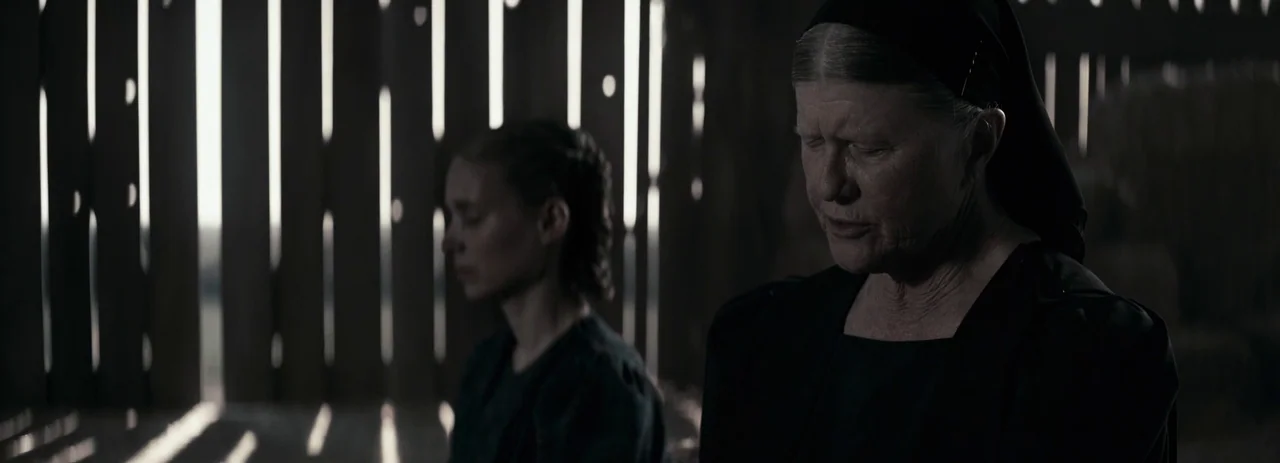 Women Talking (2022) movie still: medium — Two women stand in a dimly lit, rustic wooden building with vertical slats. They both have their eyes…