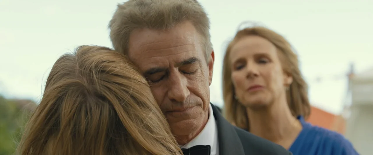 Anyone But You (2023) movie still: closeup — An outdoor gathering with neutral daylight and soft focus background. A man in a tuxedo embraces an…