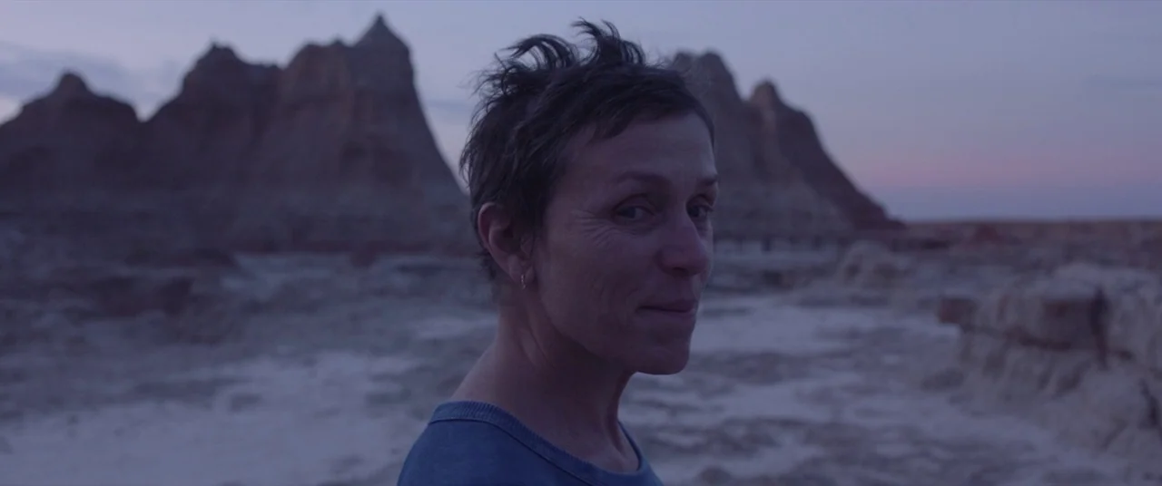 Nomadland (2020) movie still: medium — A woman stands amidst the weathered, stratified rock formations of a desert landscape at dusk. The…