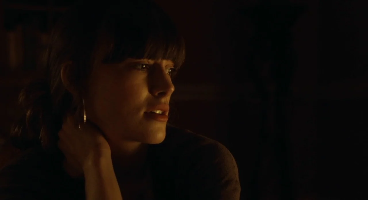 London Boulevard (2010) movie still: closeup — A dark, dimly lit room is illuminated by a single warm light source. A young woman with dark hair…