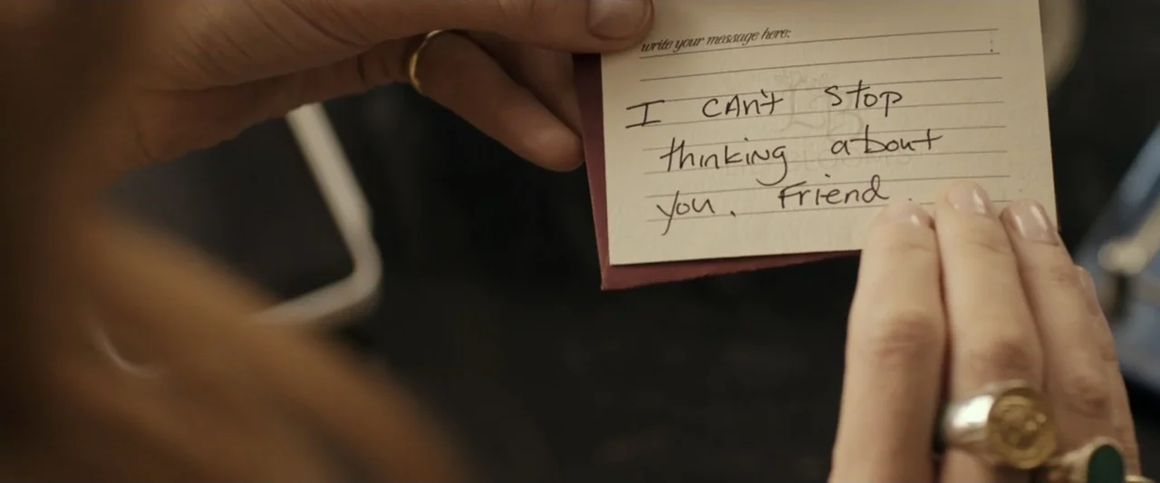 It Ends with Us (2024) movie still: closeup — Close-up view shows a hand holding a small card with handwritten text. The card rests against a dark,…