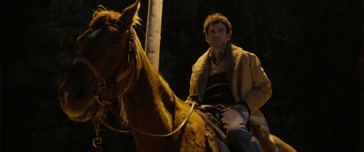 Big Bad Wolves (2013) movie still: medium — A man rides a horse at night next to a wooden utility pole. Dark trees fill the background beyond the…