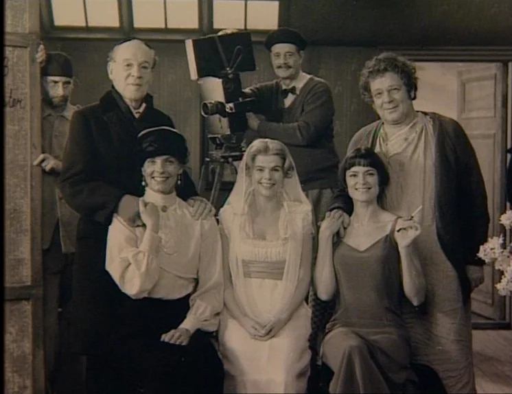 In The Presence of a Clown (1997) movie still: medium — A group of six people pose for a formal portrait inside a film studio. A vintage film camera…