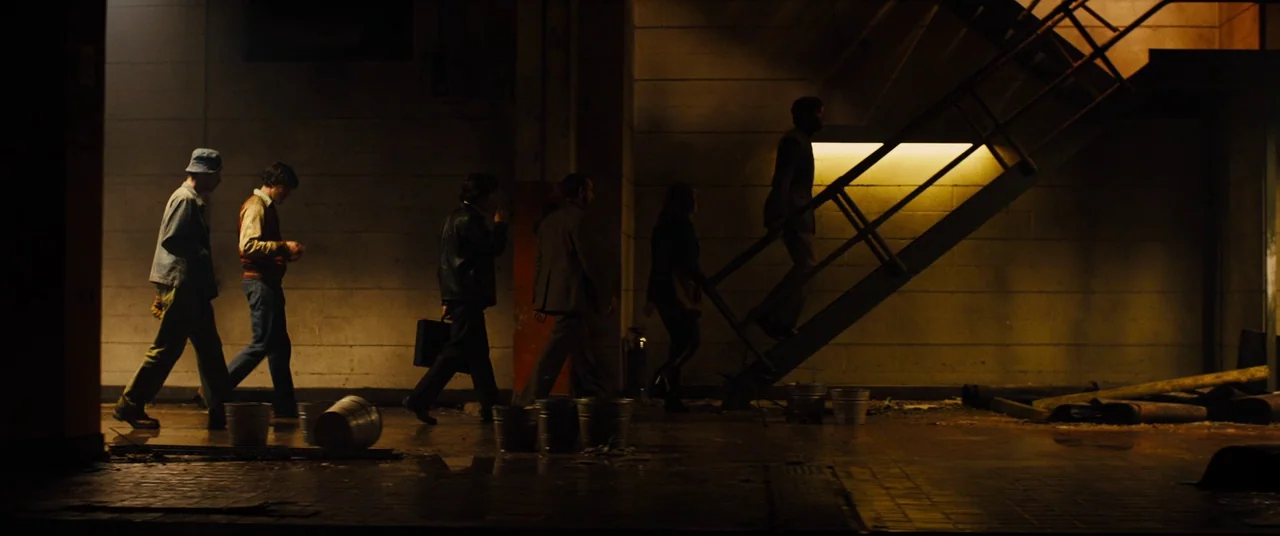 Free Fire (2016) movie still: wide — A dark subterranean space features cinder block walls and a metal staircase on the right. A small group of men…