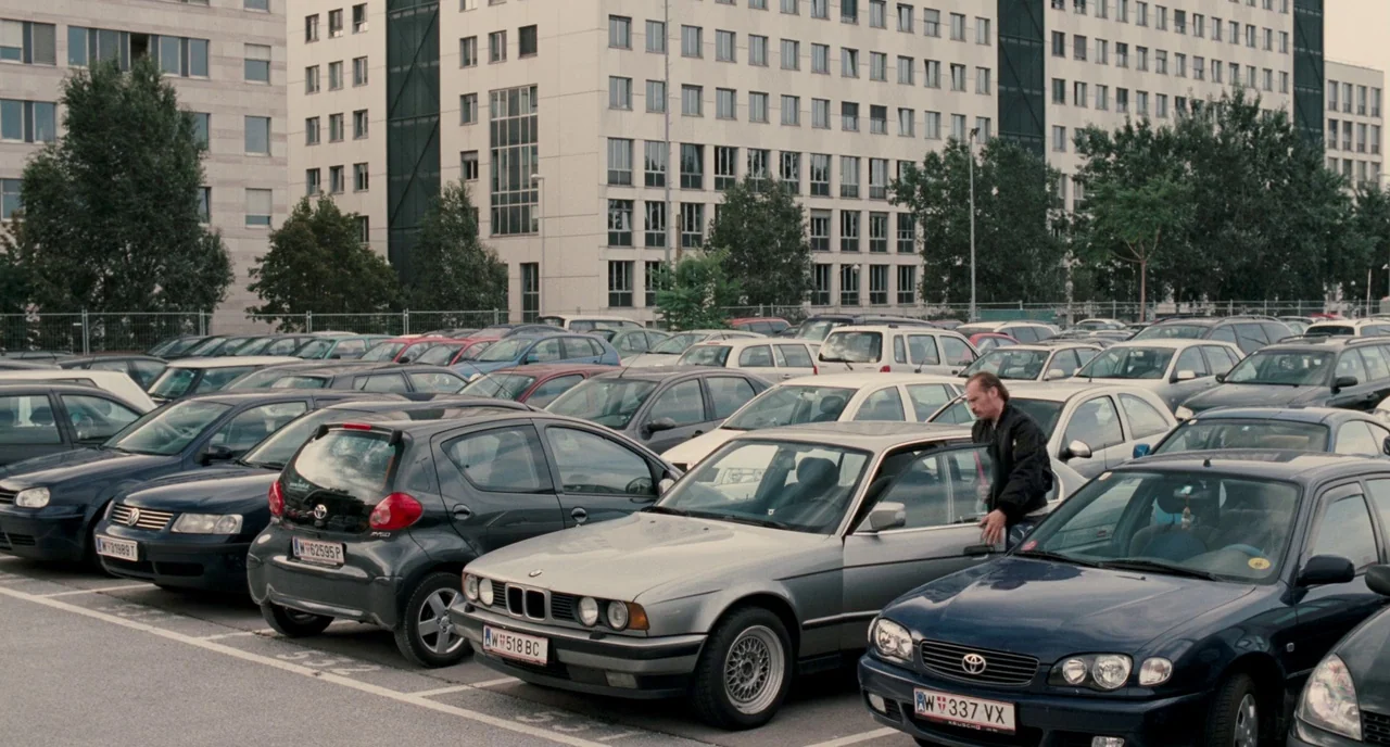 Revanche (2008) movie still: wide — A crowded asphalt parking lot features numerous cars parked in front of office buildings. A middle-aged man…