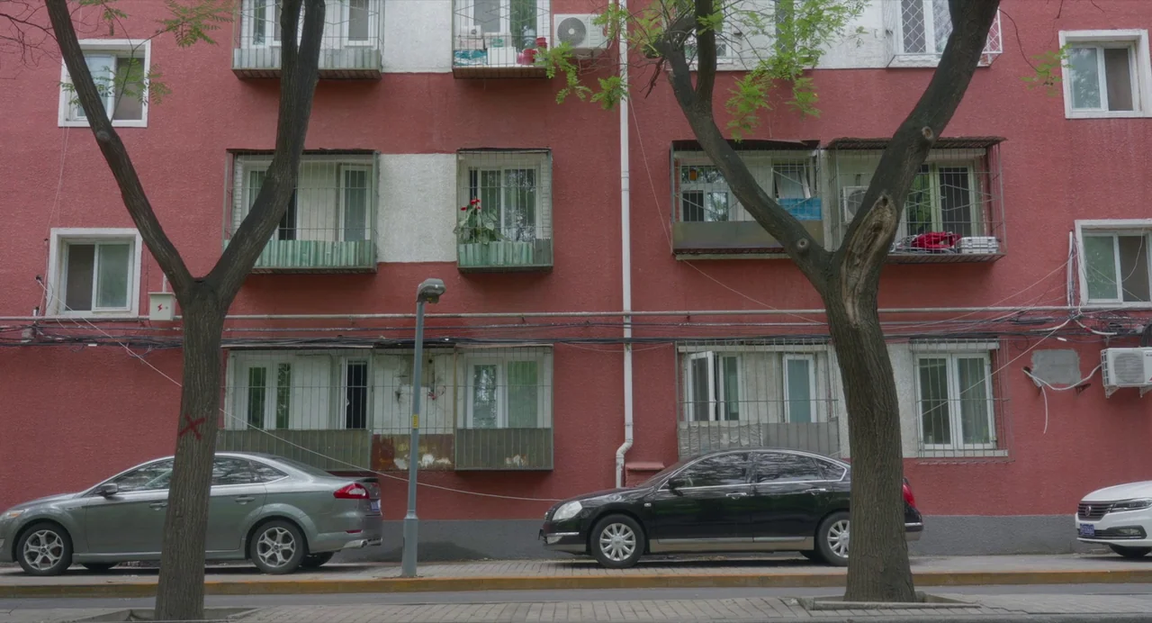 White River (2023) movie still: establishing — A red residential building wall features multiple windows covered by metal security grilles. Three…
