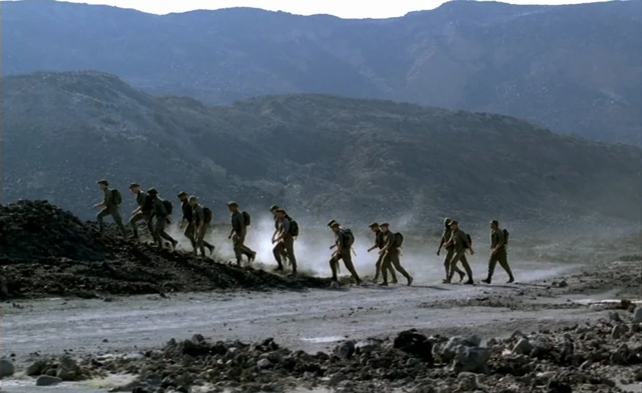 Beau Travail (1999) movie still: wide — A line of soldiers in uniform traverses a desolate, rocky desert terrain with mountains. The group moves…