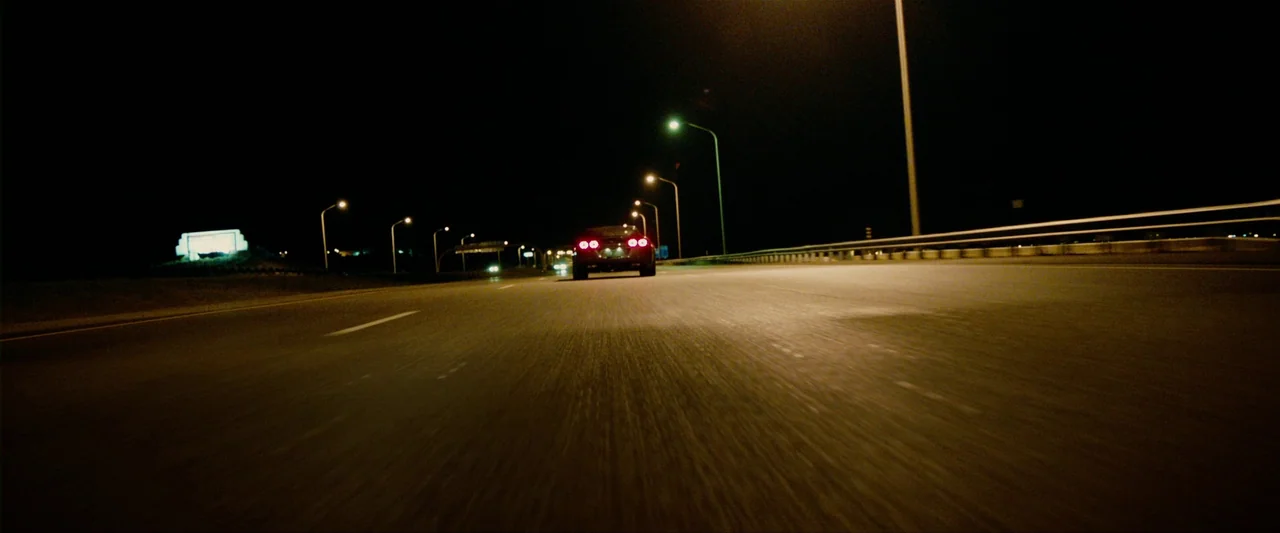 The Last Stand (2013) movie still: wide — An empty multi-lane asphalt highway stretches into the distance under darkness. A car with glowing red…