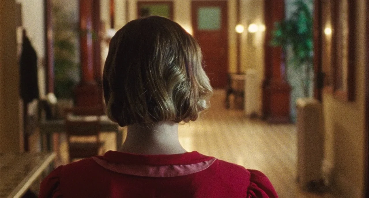 Chained For Life (2018) movie still: medium — A long interior hallway features dark wood molding and a light-colored tiled floor. A woman in a red…