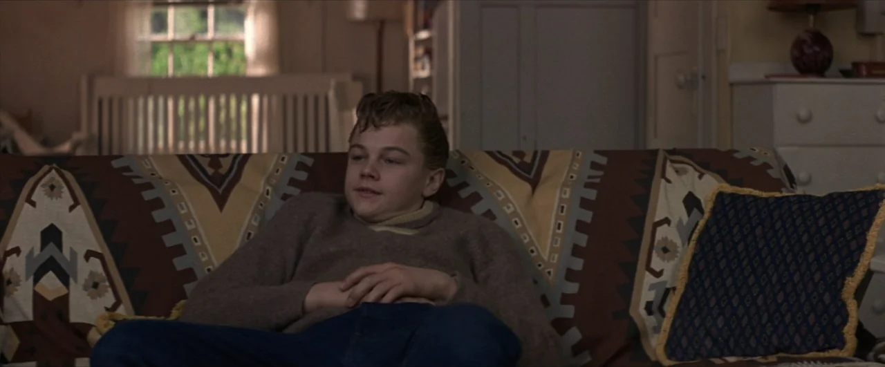 This Boy's Life (1993) movie still: medium — A domestic interior features a patterned throw over a sofa with a window in back. A teenage boy sits on…