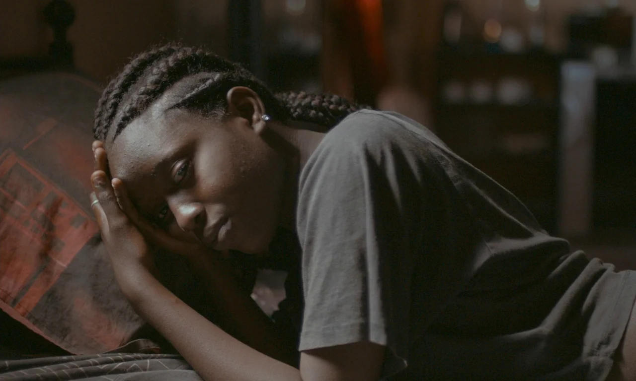 Atlantics (2019) movie still: medium — A dark bedroom with a person lying on a bed against a patterned pillow. The subject rests their head on their…