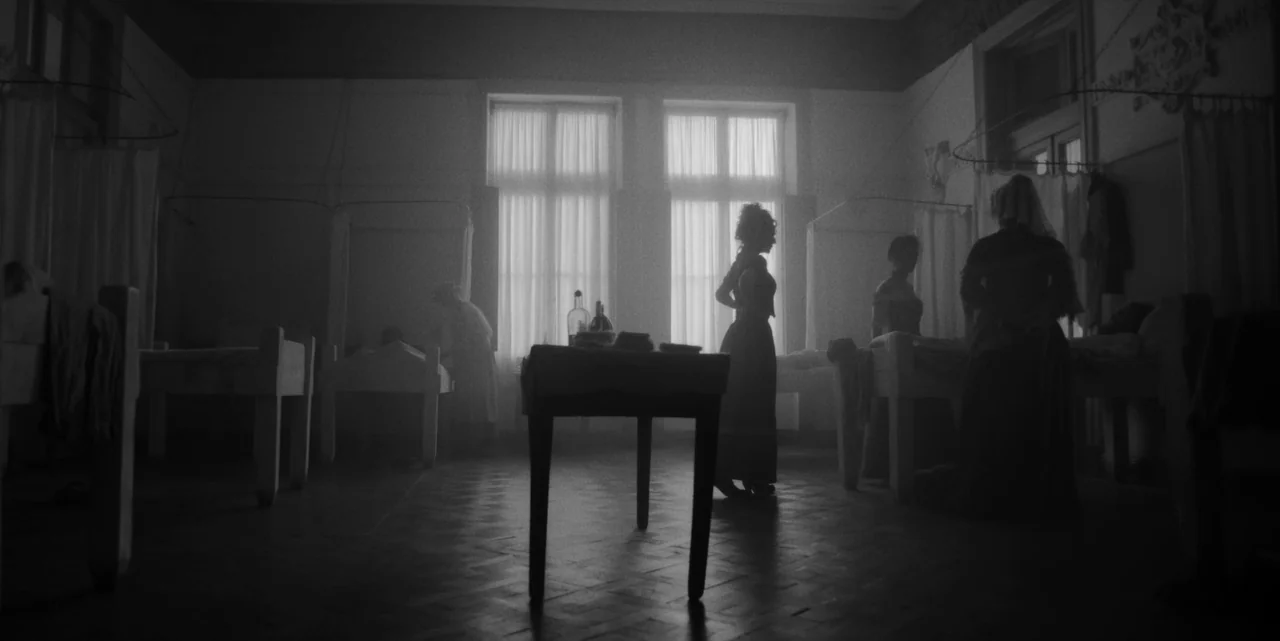 El Conde (2023) movie still: wide — An early 20th-century hospital ward features several iron-framed beds arranged in rows. Nurses in long dresses…