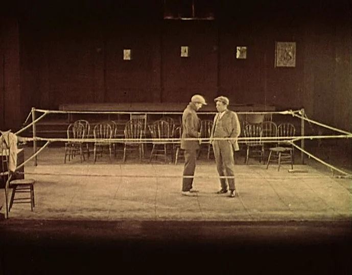 Broken Blossoms (1919) movie still: wide — An indoor space features a small rope-lined ring surrounded by several wooden chairs. Two men wearing…