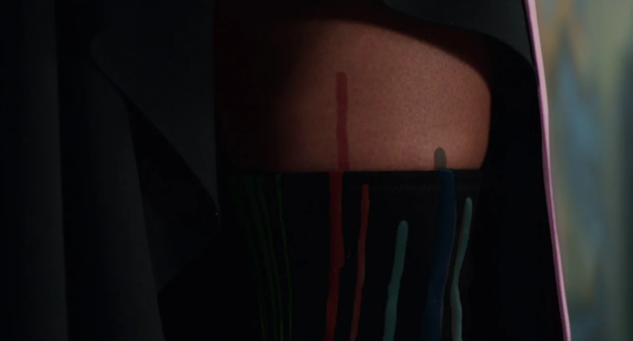 Velvet Buzzsaw (2019) movie still: closeup — A close view of a person's thigh against dark fabric. Multiple colored paint streaks run vertically…