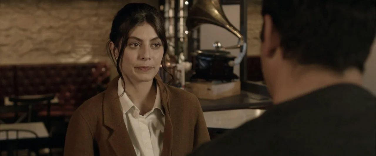 About Us (2017) movie still: medium — An interior cafe setting features a tufted leather booth and a vintage gramophone. A young woman sits at a…