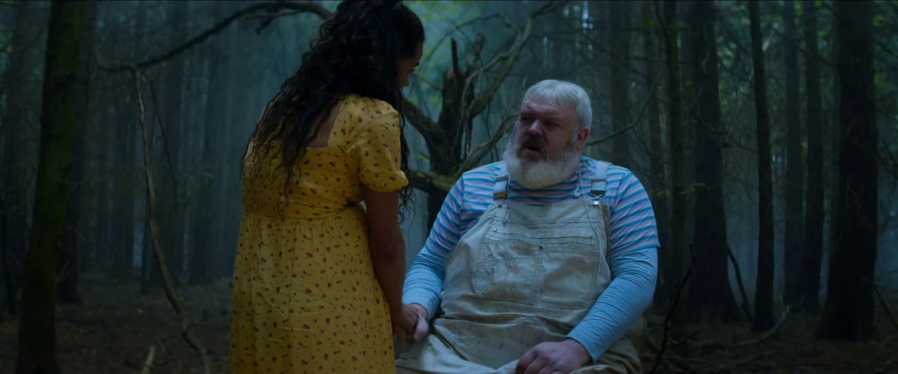 Unwelcome (2022) movie still: medium — A dense, misty forest filled with tall tree trunks and bare, tangled branches. A young woman in a yellow…