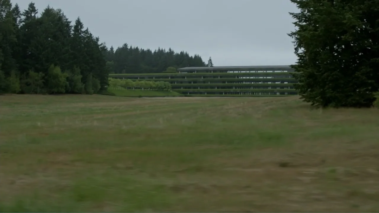 Trees and Other Entanglements (2023) movie still: establishing — A low-slung, multi-tiered office building with green landscaping sits behind a…