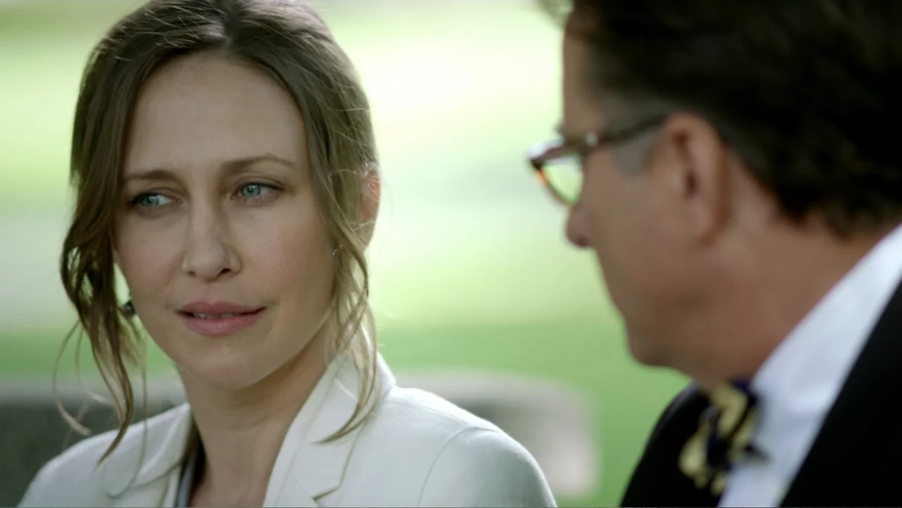 At Middleton (2013) movie still: medium — An outdoor park setting with a blurred green background. A woman in a white blazer gazes toward a man…