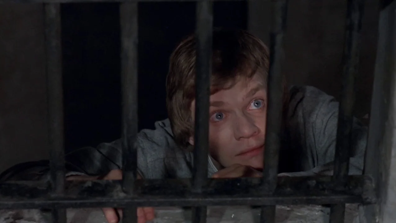 O Lucky Man! (1973) movie still: closeup — A man looks through the iron bars of a dark stone enclosure. The subject presses his hands and face…