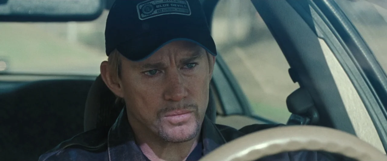 Roofman (2025) movie still: closeup — A man drives a car during the daylight hours with both hands on the wheel. The view through the windshield…