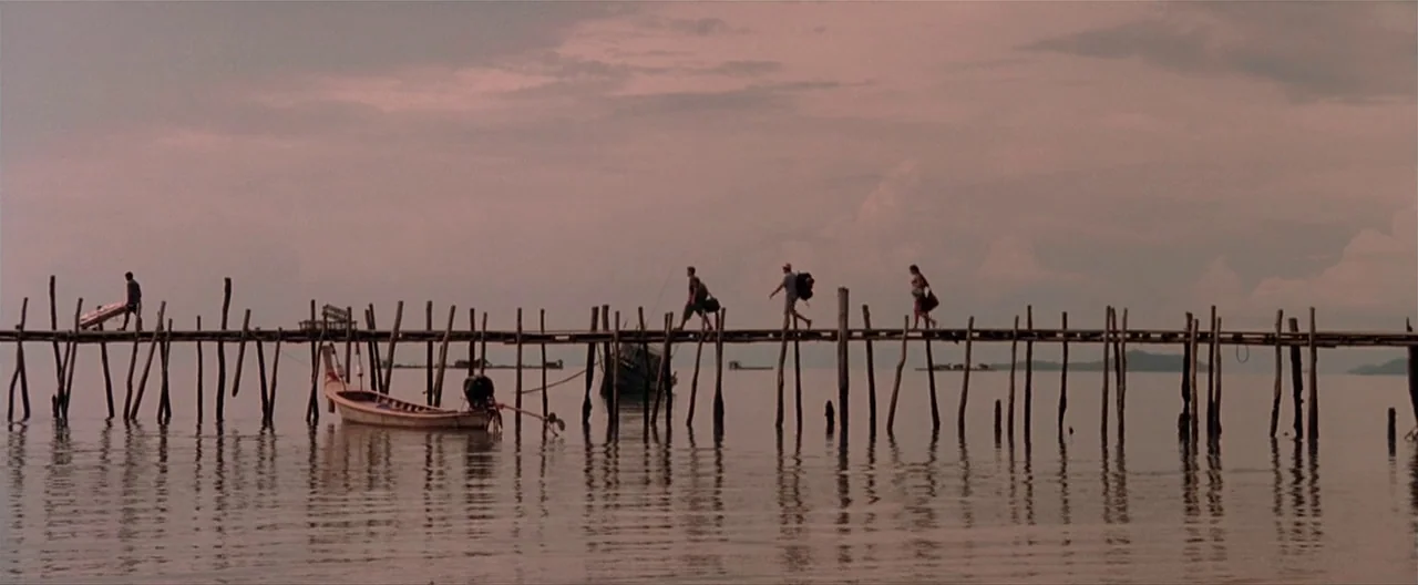 The Beach (2000) movie still: wide — A long wooden pier extends over flat water under a pale pink sky. Four travelers walk across the pier carrying…