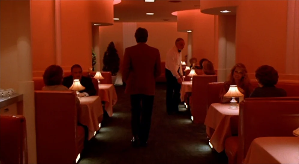 American Gigolo (1980) movie still: wide — A dimly lit restaurant features orange booths, white tablecloths, and small lamps on tables. A man in a…