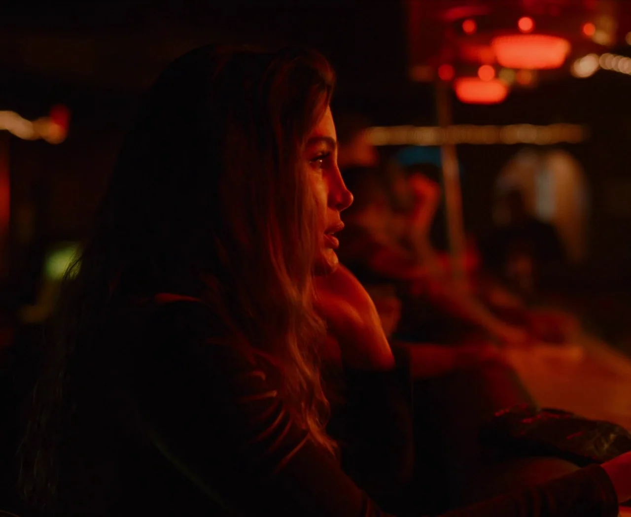 Monica (2022) movie still: medium — A dark bar interior is illuminated primarily by red neon lights overhead. A woman with long brown hair sits at a…