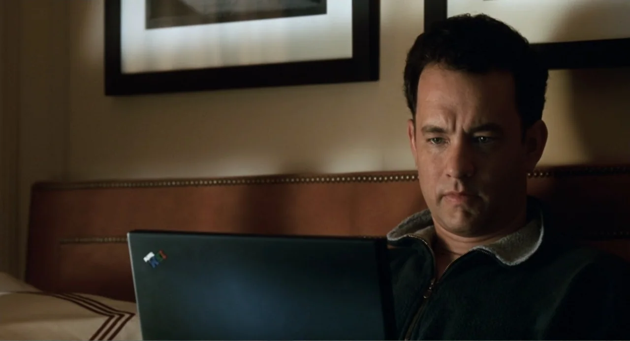 You've Got Mail (1998) movie still: medium — A man sits in bed in a dark room with a padded headboard behind him. He focuses intently on a black…