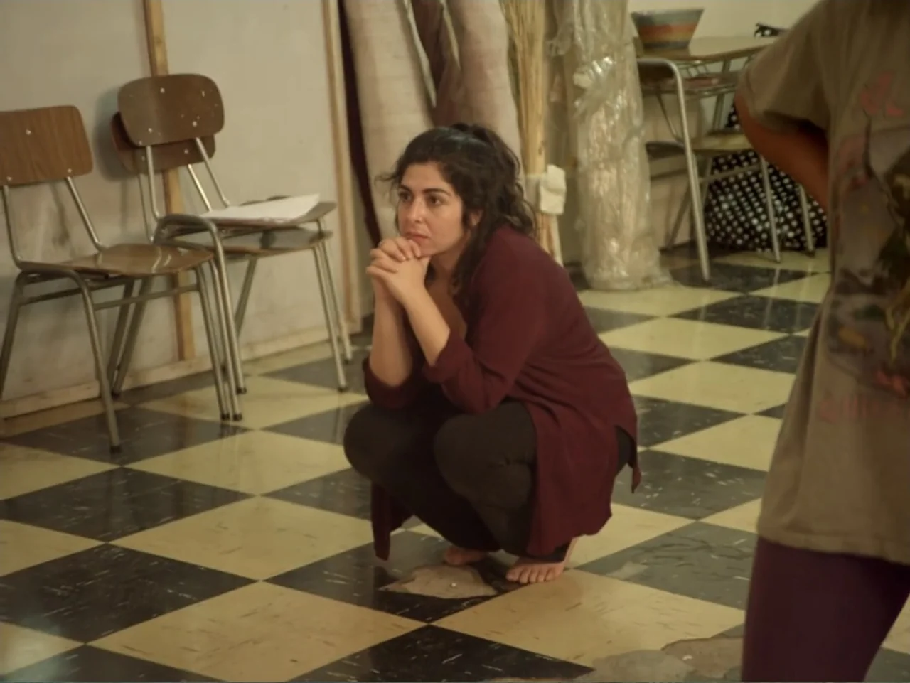 History and Geography (2024) movie still: medium — A room with a black and white checkered floor and wooden chairs at the side. A woman in a dark…