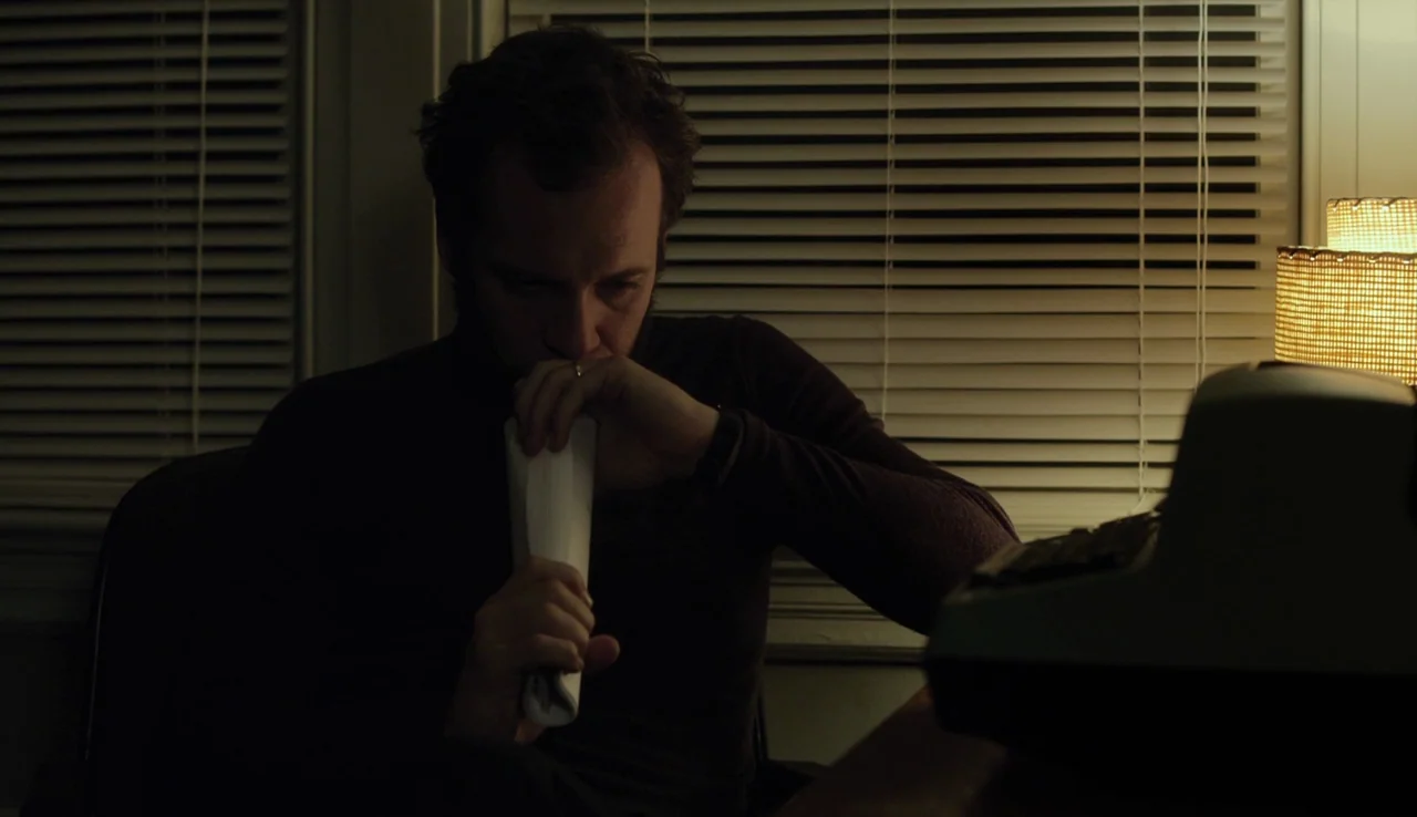 Experimenter (2015) movie still: medium — A dark room features a man sitting before a desk with horizontal window blinds. He holds a rolled document…
