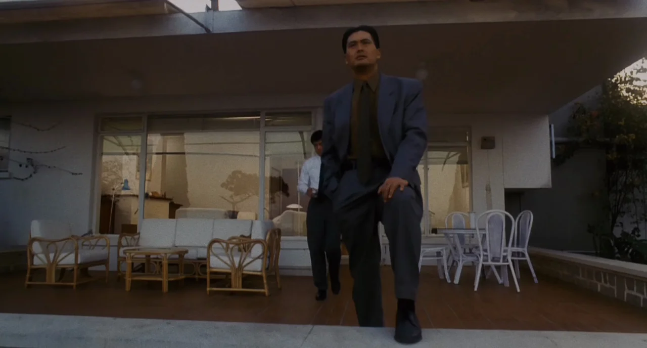The Killer (1989) movie still: fullbody — A modern patio features glass sliding doors and white wicker outdoor furniture on tiles. Two men in formal…
