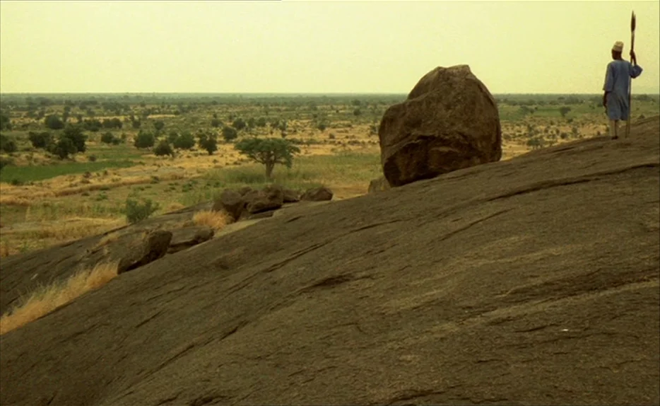 Chocolat (1988) movie still: wide — A vast, dry landscape extends toward the horizon under a pale, hazy sky. A lone figure stands on a prominent…