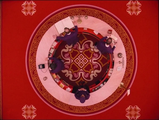 The Grand Budapest Hotel (2014) movie still: establishing — A circular concierge desk sits centrally on a red patterned carpet with ornate geometric…