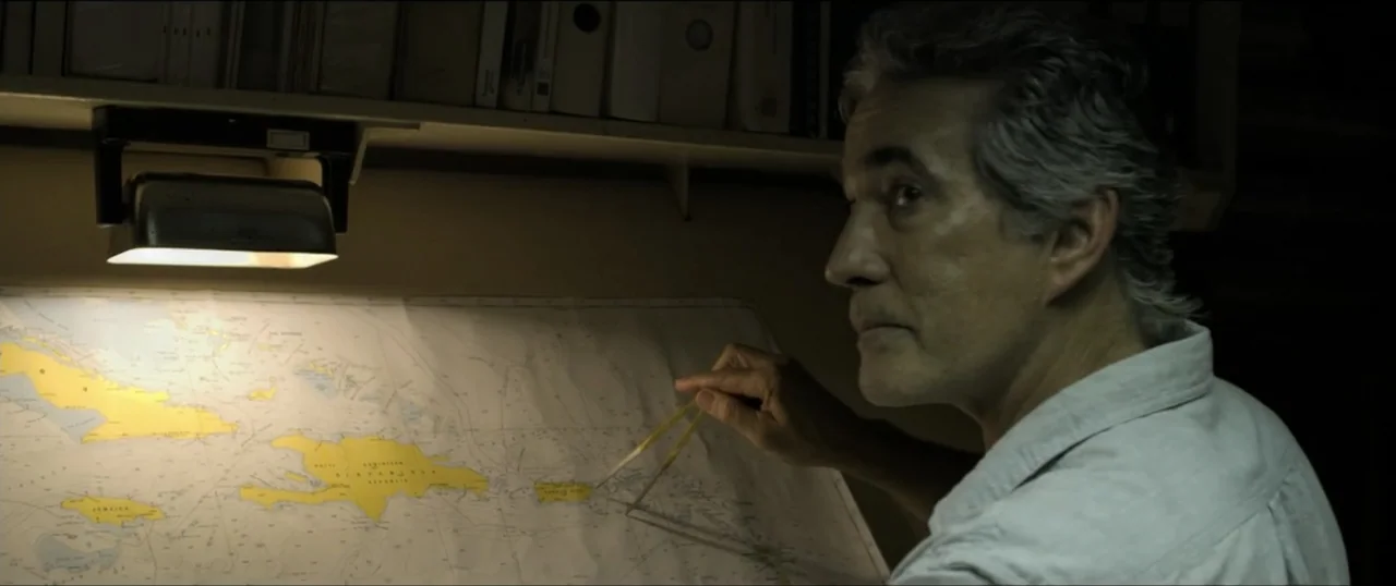 Primal (1994) movie still: medium — A dimly lit room contains a desk with a large Caribbean map spread across it. A middle-aged man in a light grey…